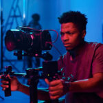Cinematographer operating a professional camera rig on a film set with pink and blue lighting.